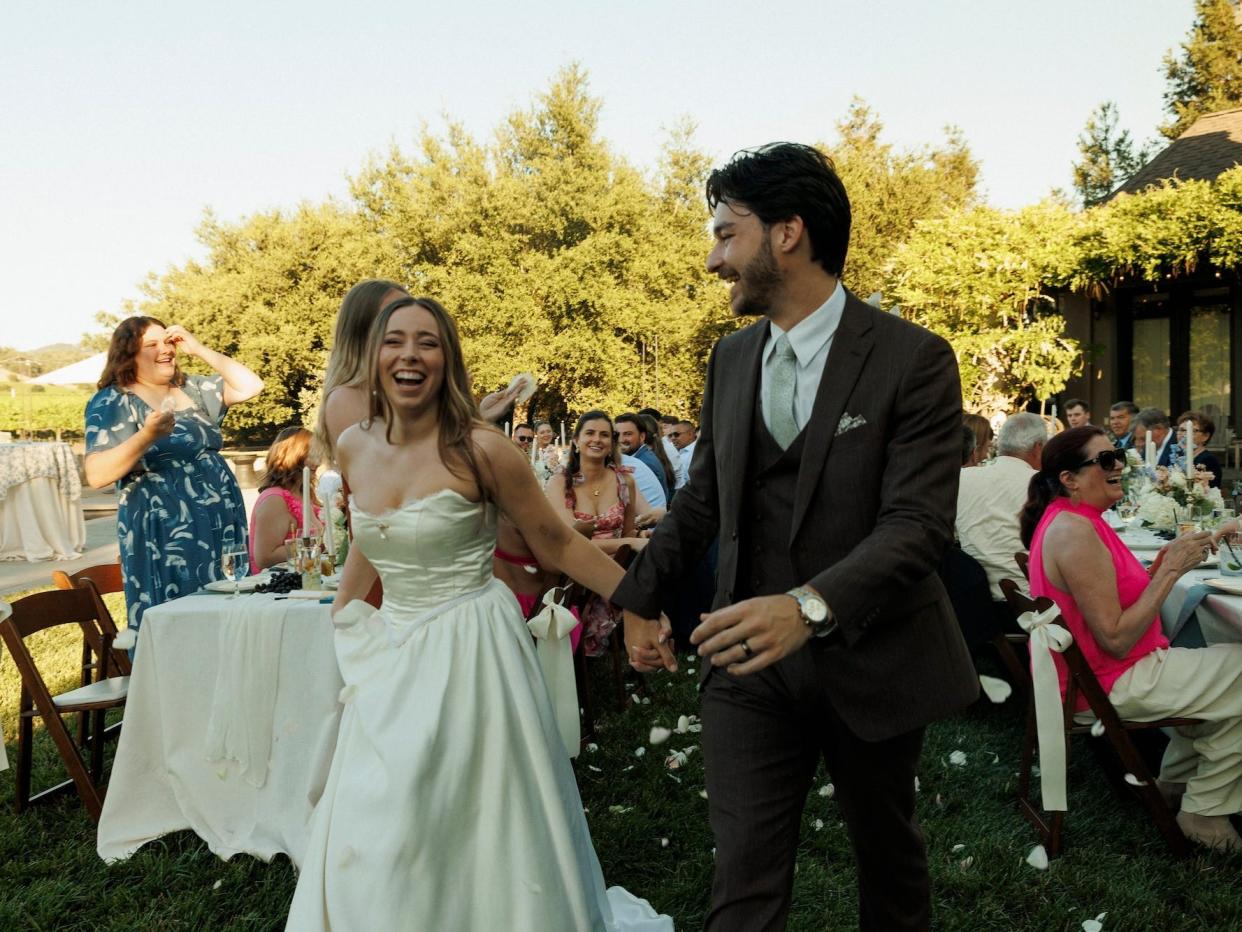 A bride and groom hold hands as they walk into their wedding reception with flower petals around them.