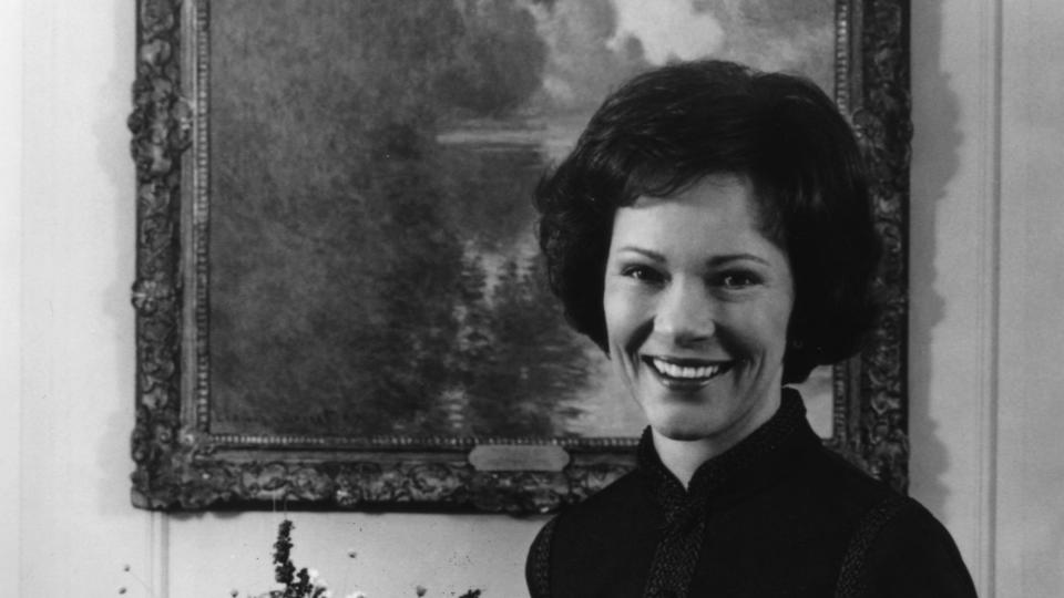 rosalynn carter smiles and stands behind a chair and in front of a large floral bouquet and painting