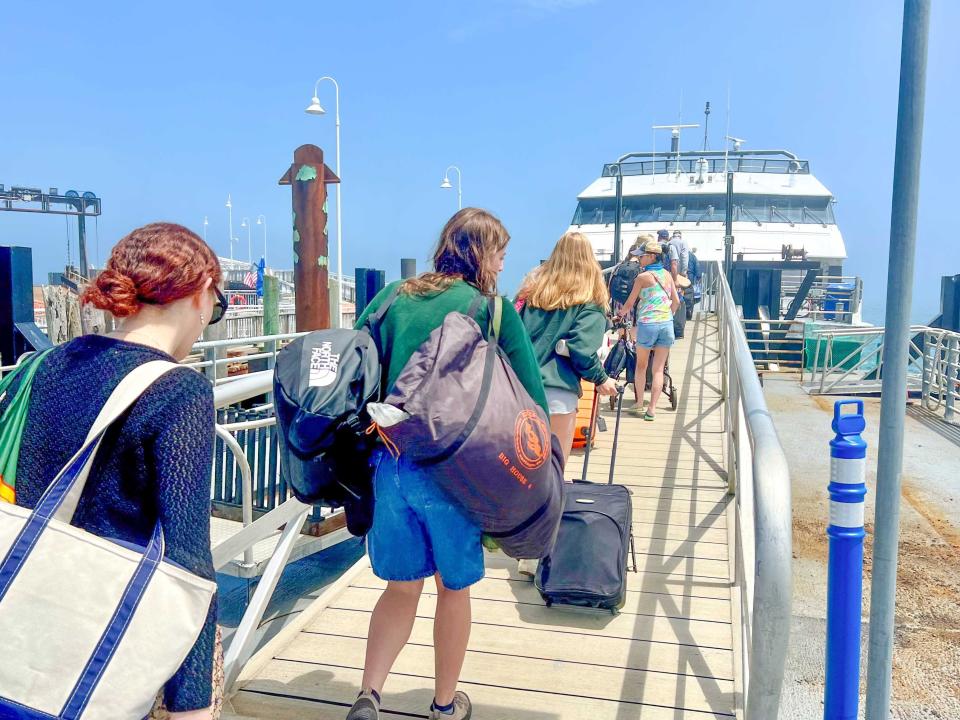 Several passengers with bags and totes are lined up on a dock and boarding a large white ferry. Two women with a blue cardigan and white tote bag and a green sweatshirt and two duffle bags are in the foreground.