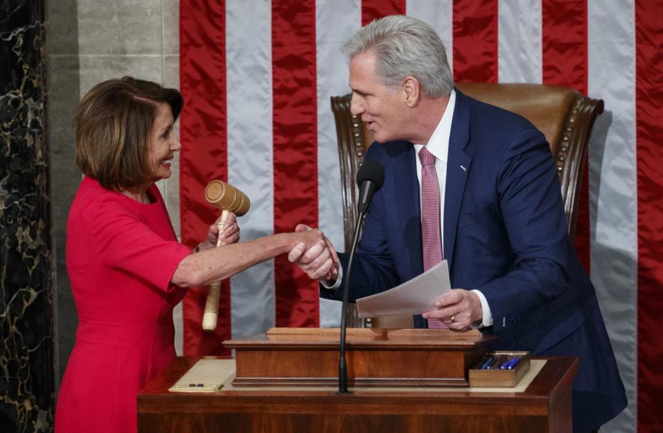 A woman in a red dress with a gavel shakes hands with a man in a suit in front of a U.S. flag.