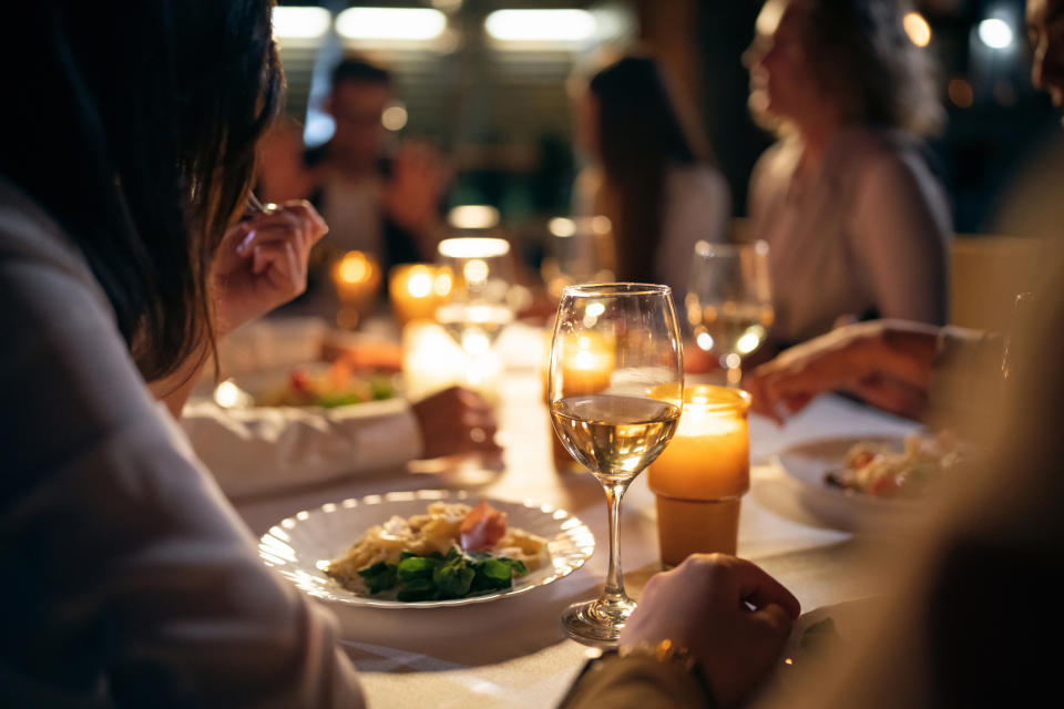 People are gathered around a dining table with plates of food and drinks, conversing in a dimly lit, candle-lit setting. Names of persons unknown