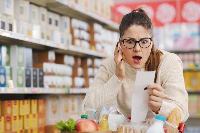 A shocked woman in a supermarket holding a receipt while pushing a shopping cart full of various items