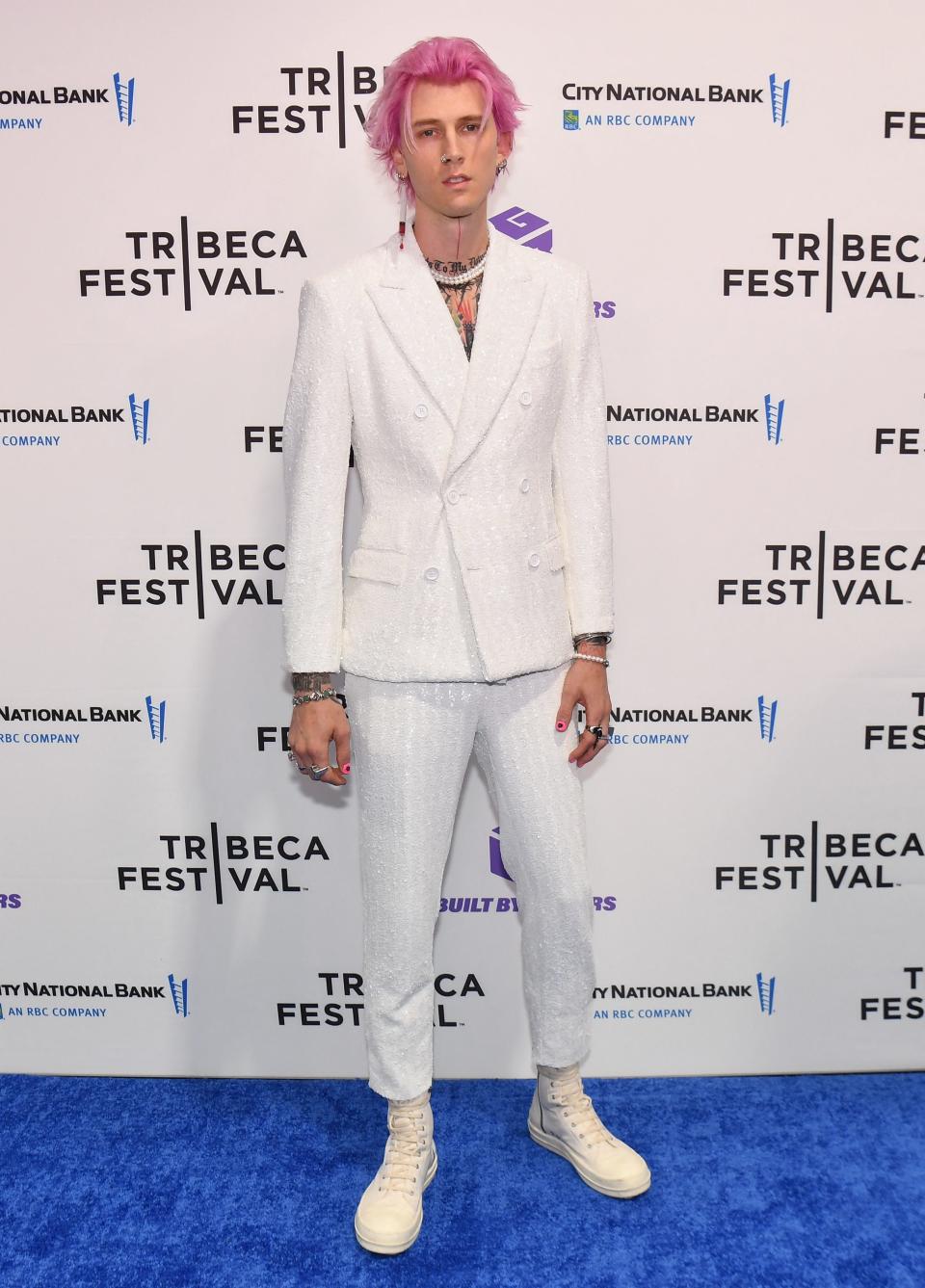 machine gun kelly in a sequined white suit, standing on a red carpet. he has pink hair pushed back from his forehead and an earring shaped like a syringe dangling from his right ear