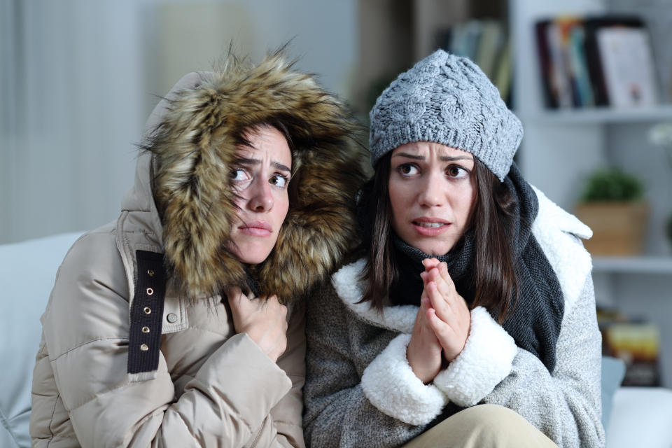 Two women are sitting indoors, dressed warmly in winter coats and hats, looking worried