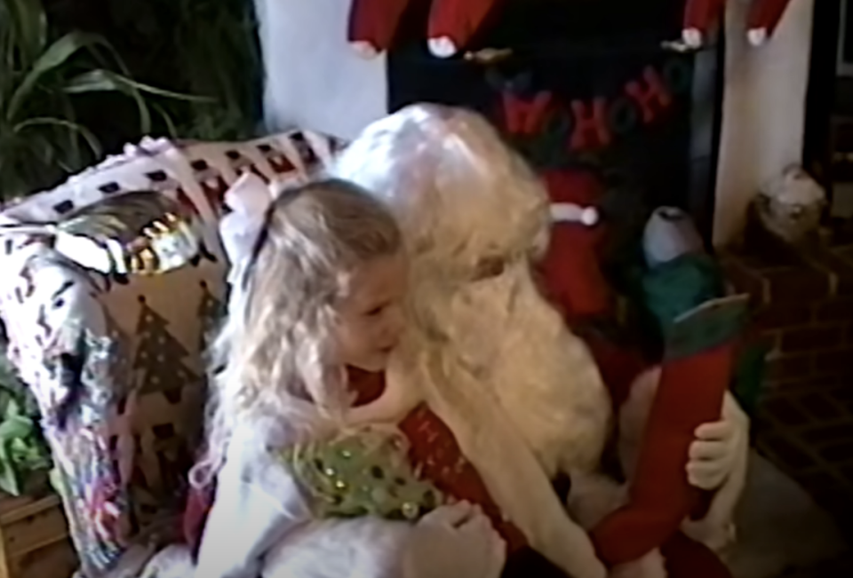 A child sits on Santa's lap by a fireplace with stockings, holding a stocking and conversing