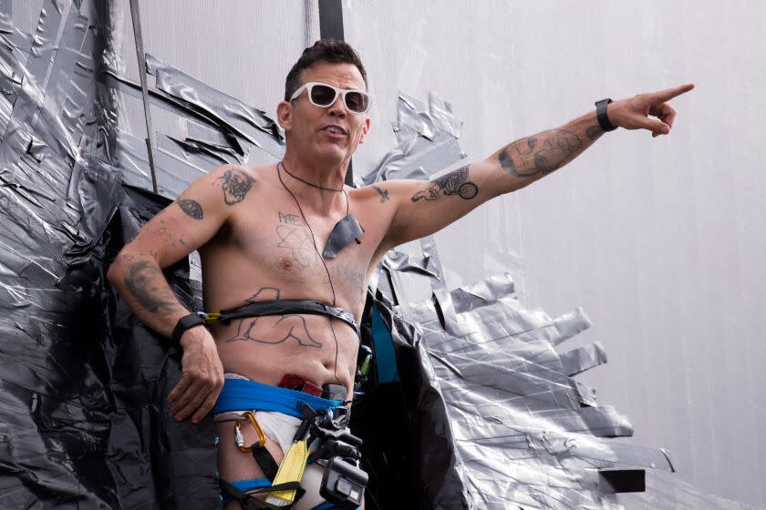 Steve-O is pointing while being tapped to a billboard and is shirtless wearing sunglasses
