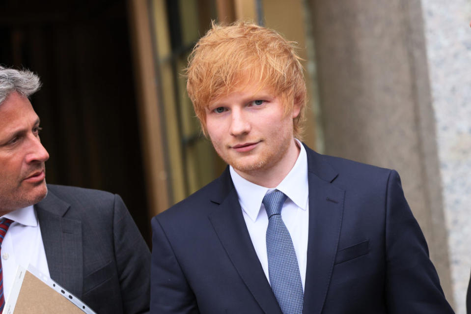 Ed Sheeran and an unidentified man dressed in suits, standing outside a building. Ed Sheeran is wearing a navy suit with a white shirt and gray tie