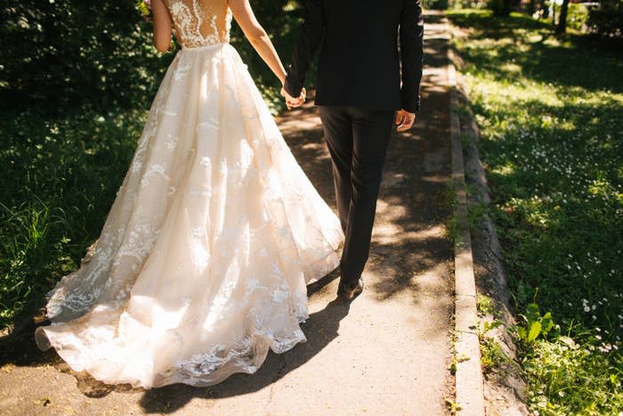 Bride in an elegant, lace wedding gown and groom in a tailored black suit holding hands while walking down a park pathway