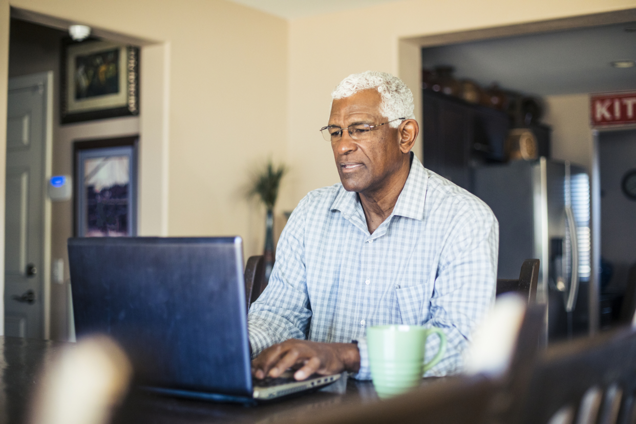 Senior man on his laptop at a desk in his living room, with an aqua coffee cup and a beige corner wall behind him with the kitchen blurred in the background