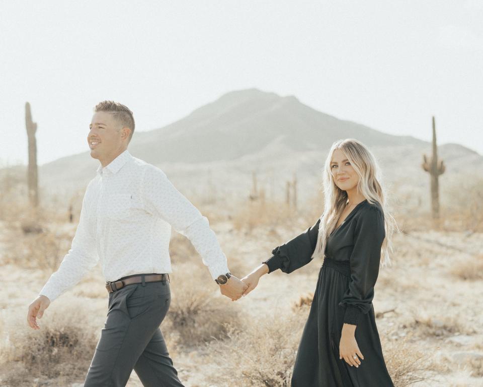 A man and woman hold hands and walk to the left of the frame in the desert.