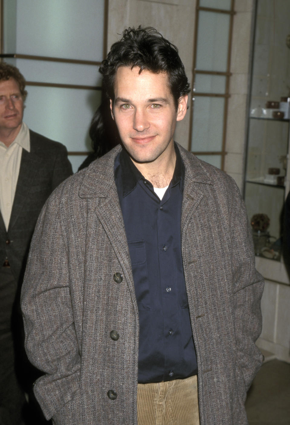 Paul Rudd stands indoors wearing a dark shirt, brown trousers, and a gray overcoat, with hands in pockets. Another man is visible in the background