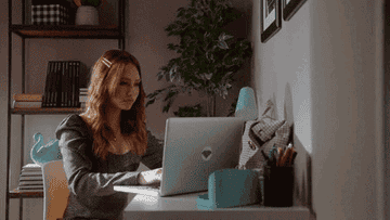 A woman sits at a desk working on a laptop in a home office with shelves, books, and plants in the background