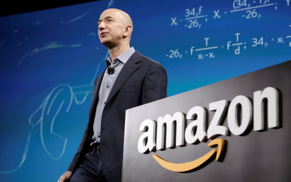 Jeff Bezos, a pale-skinned man with no hear wearing a business suit, is seen from a low angle as he speaks in front of a blue backdrop scrawled with equations from an Amazon stage - Jason Redmond/Reuters