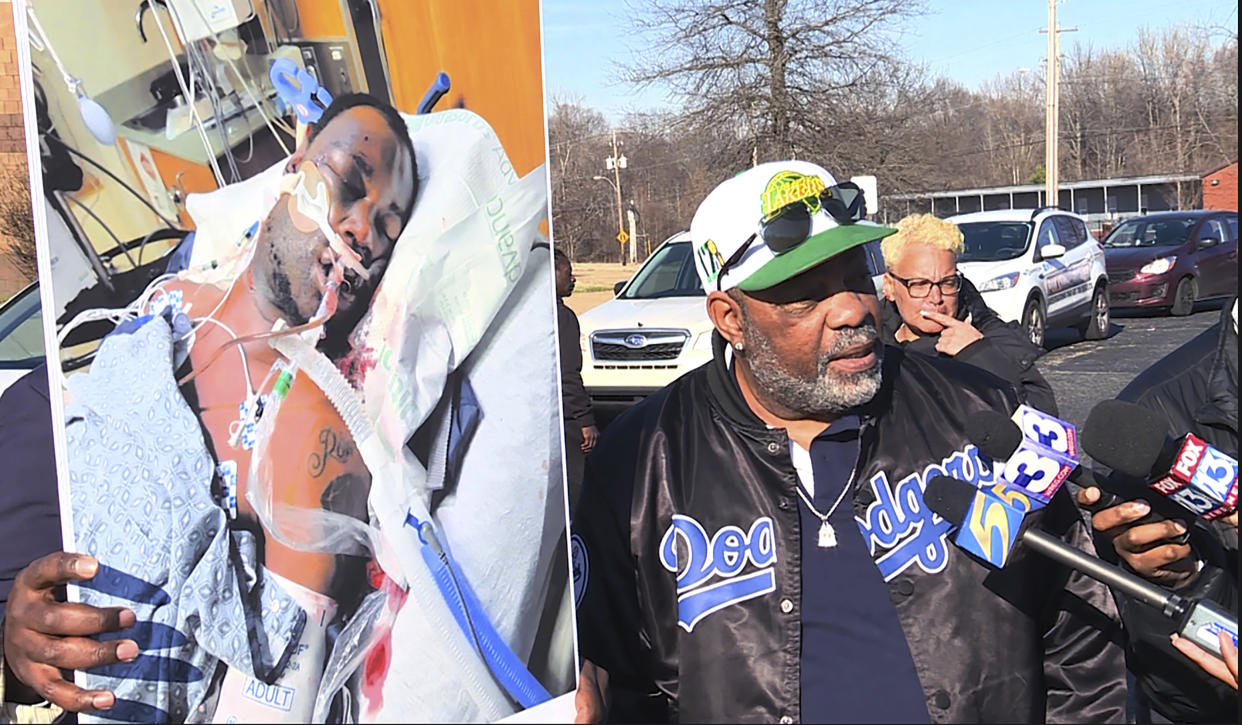 Rodney Wells, outside, surrounded by cars, protesters and media microphones, holds a poster showing Tyre Nichols, his face covered with dark bruises, in a hospital bed on life support.