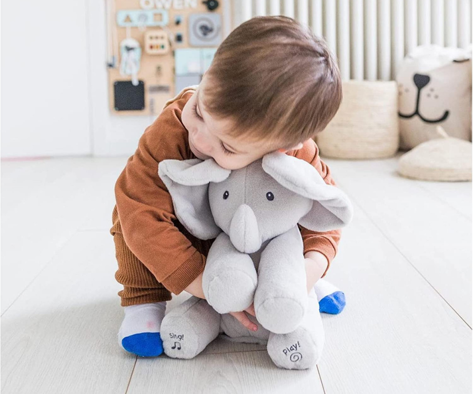 A little boy with brown hair crouches and cuddles a grey soft toy elephant on a white floor with pale cream toys in the background.