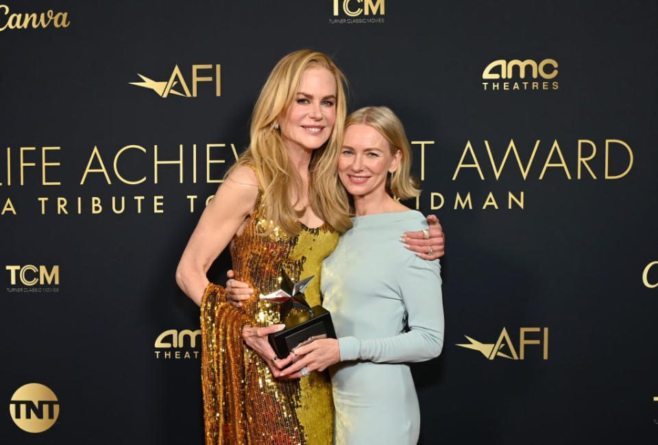 Two women posing with an award, one in a gold sequined gown and the other in a pastel blue dress