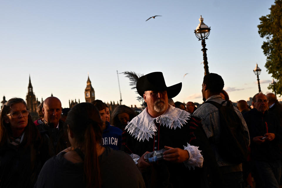 A man wearing a feathered hat and period collar stands in the queue