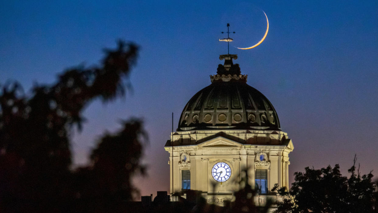  A thin crescent moon hangs in a dark blue sky, fading to a dull orange near the horizon, behind a stone, iron-domed, courthouse with a fish weather vein mounted at its peak. the silhouette of tree branches intrude from the left. 