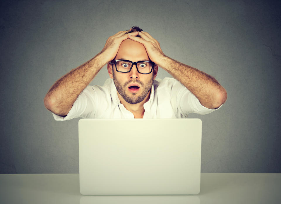 A white man wearing glasses and sitting at a table appearing to panic while staring at a laptop computer.