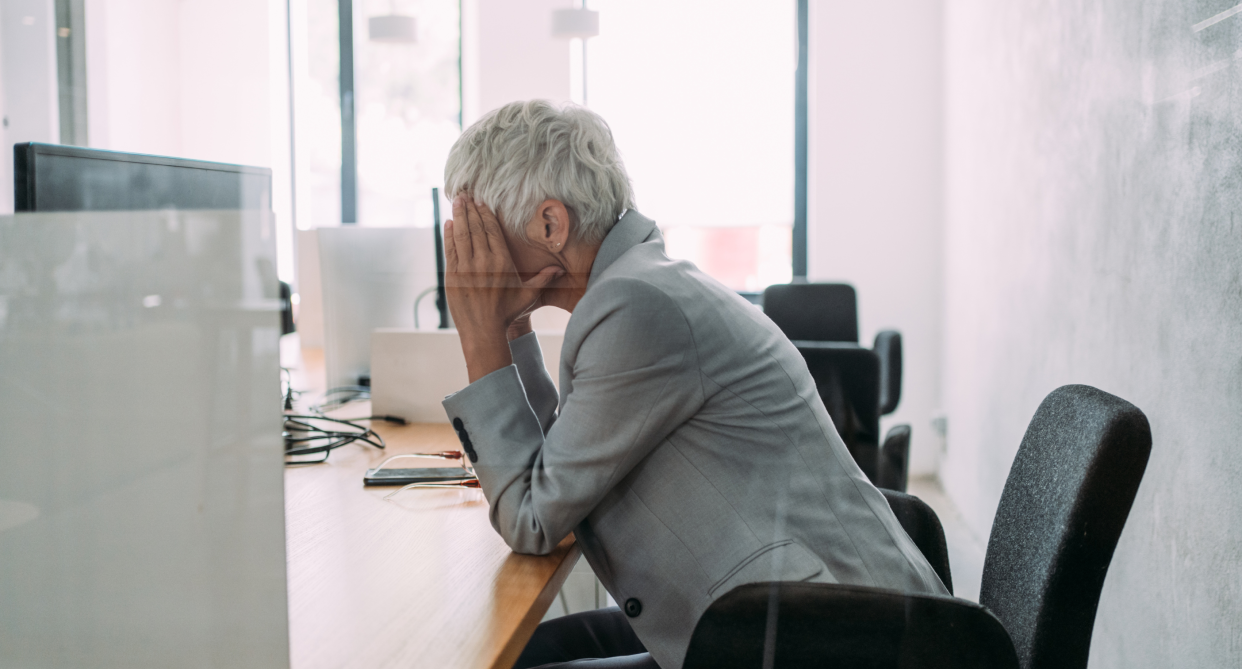 menopause, woman with short grey hair wearing grey blazer sitting in office chair with head in hands sitting at office desk