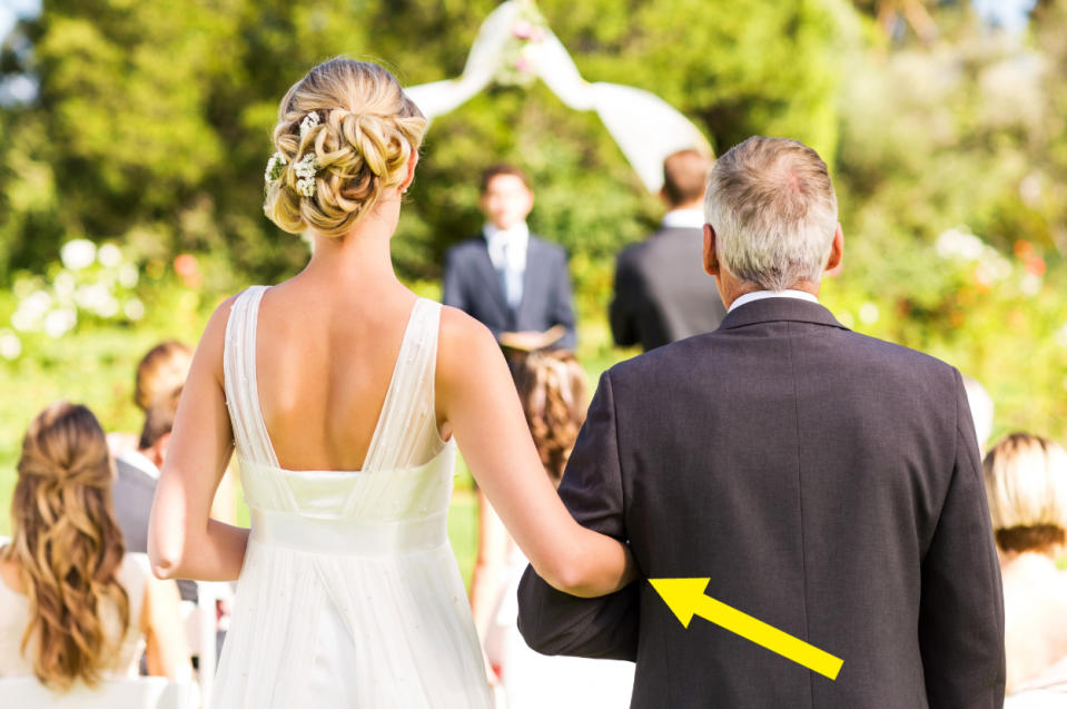 A bride in a wedding dress walks down the aisle with a man in a suit, likely her father, in an outdoor ceremony setting