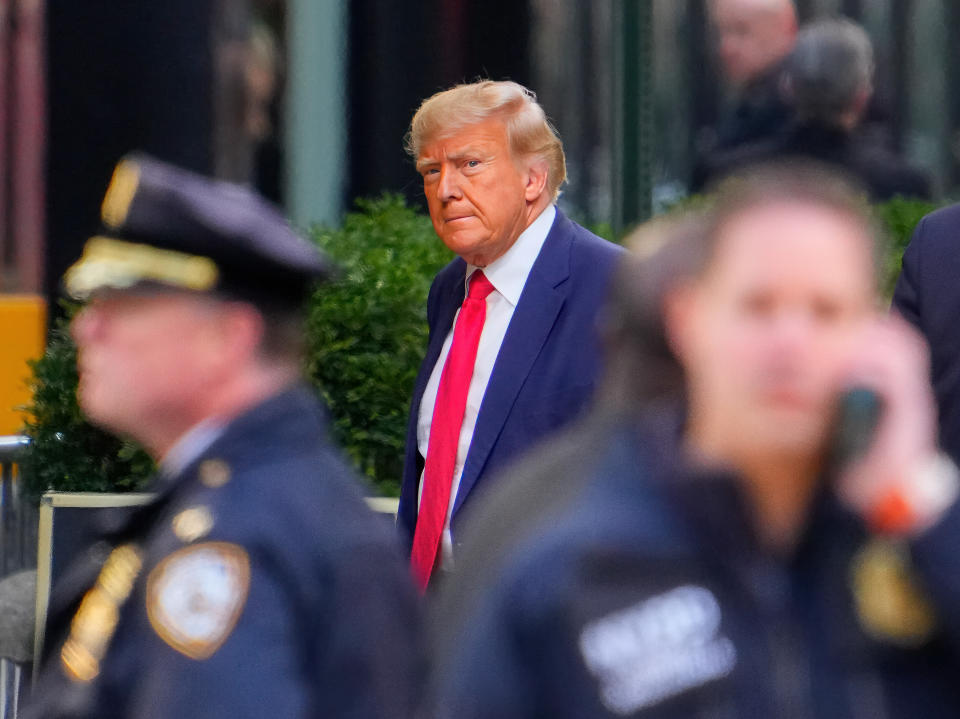 Biting his lip,  Donald Trump, in blue suit and red tie, arrives at Trump Tower, with a guard in uniform in front of him.