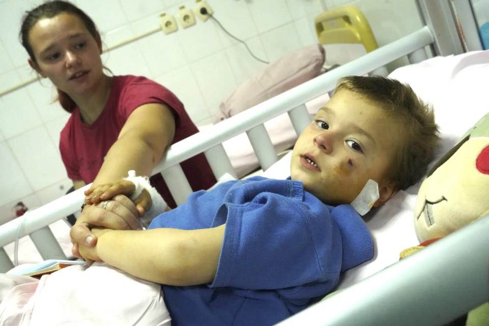 A boy in a blue shirt, with a bruise on his cheek and lying in a hospital bed, holds the hand of a woman in a red T-shirt