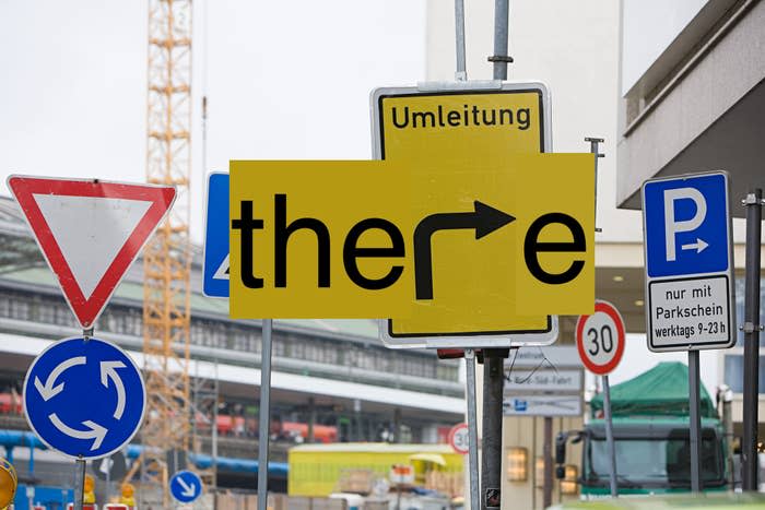 Road traffic signs including detour (Umleitung in German), parking and turn signs form the word "there." Busy street background with construction crane visible