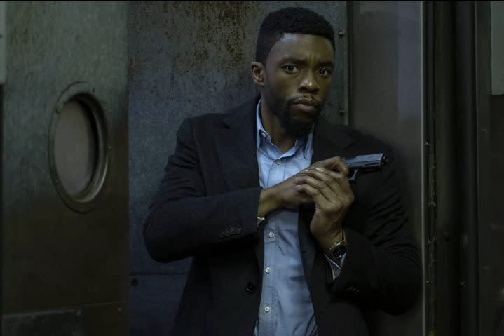 Chadwick Boseman holds a gun and stands up against a wall.