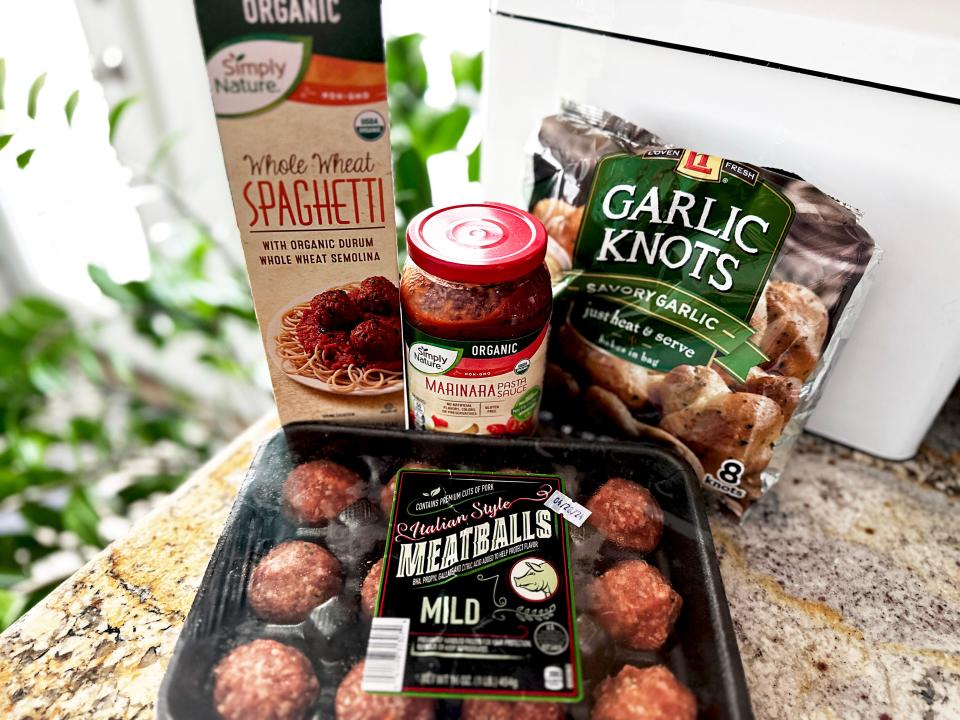 A black package containing meatballs, a bag with images of garlic knots, a jar of marinara sauce, and a green and tan box of pasta on a stone countertop