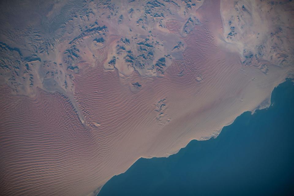 red desert sands with distant mountains against deep blue ocean