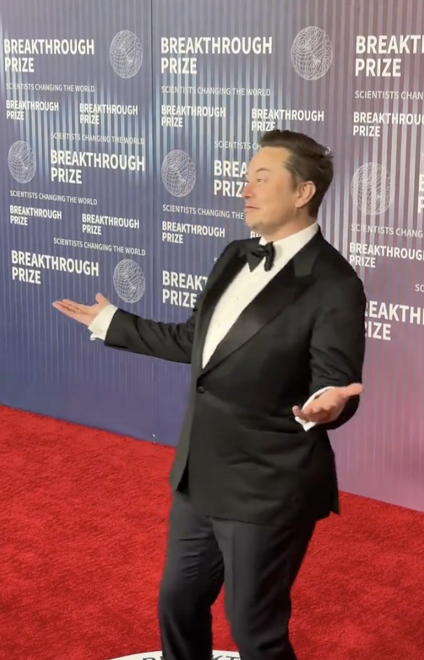 Elon Musk in a tuxedo gesturing, standing in front of a backdrop with 'Breakthrough Prize' logos