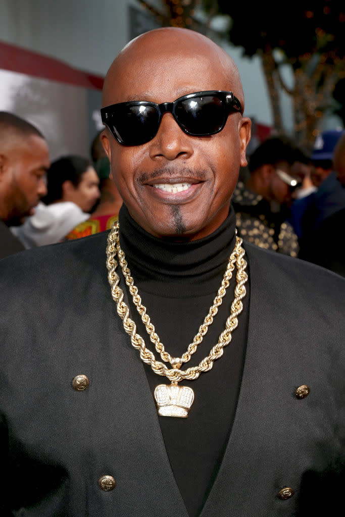 MC Hammer in sunglasses with gold chain necklaces and black jacket smiling at the camera