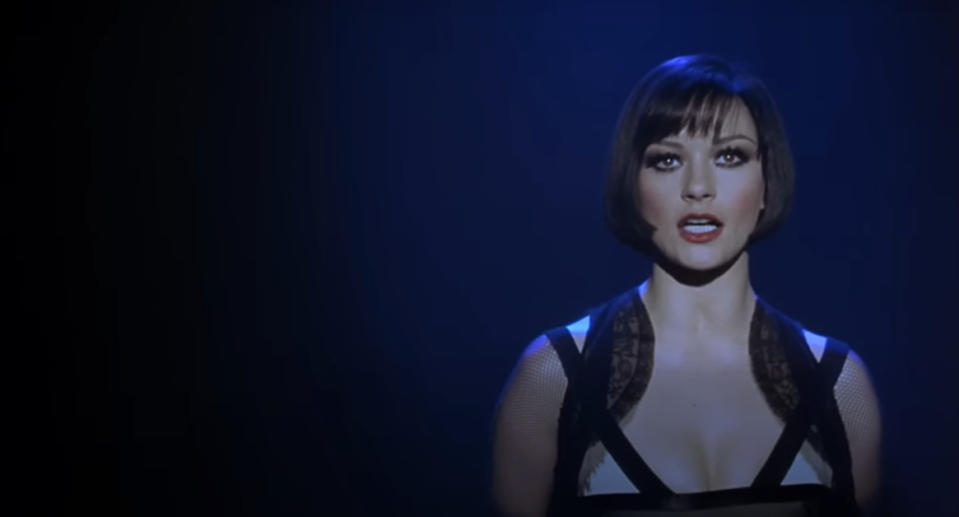 Catherine Zeta-Jones as Velma Kelly is singing passionately. She's wearing a black strappy dress with lace details.