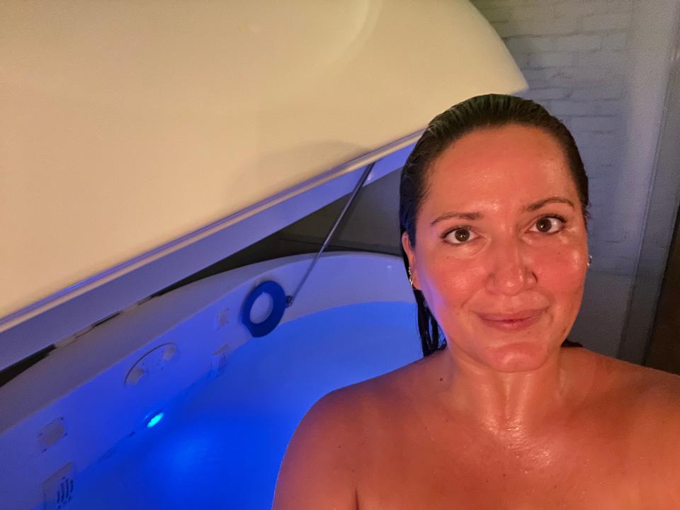 Selfie photo of Lola Méndez from the shoulders up, standing in front of a sensory deprivation tank. Her dark brown hair is wet and slicked back. She has dark brown eyes and smiles slightly.