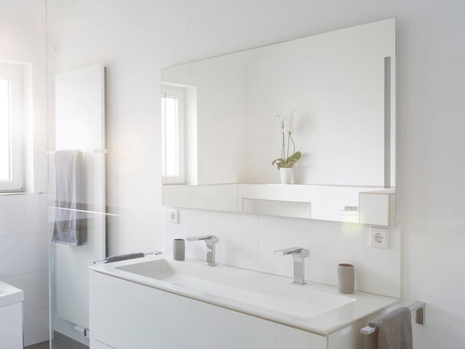 An all-white bathroom with a rectangular sink, a large mirror reflecting a flower, and clear-glass shower doors