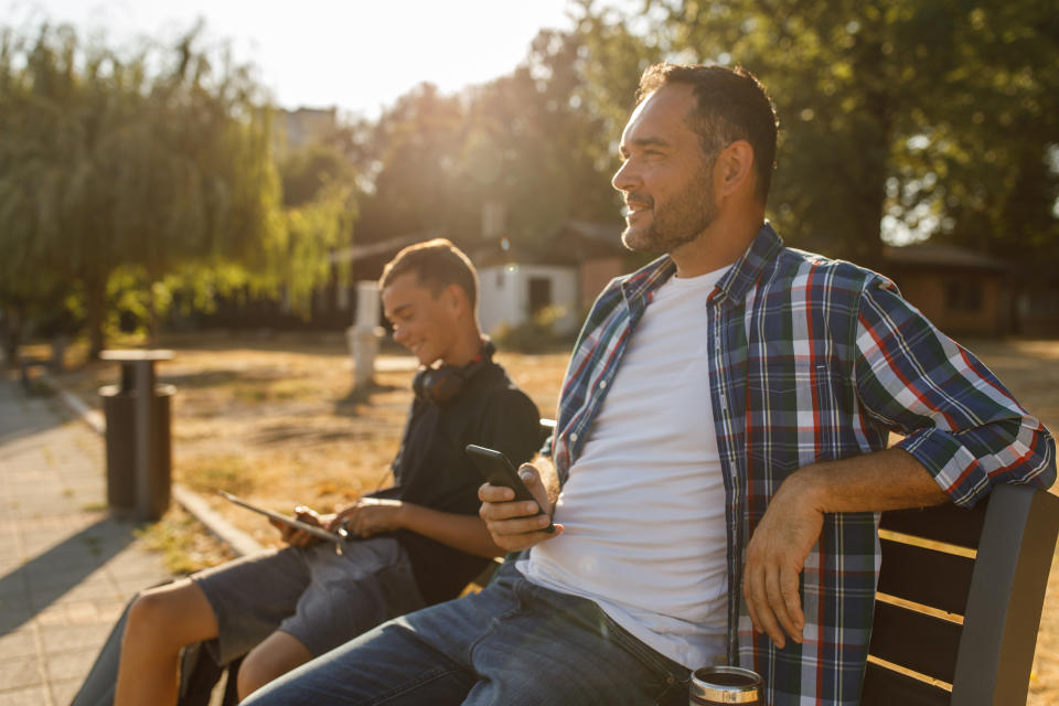 Teenage boy and his father relaxing in sunshine on the street bench. Father is using smart phone while his son is playing video games via digital tablet.