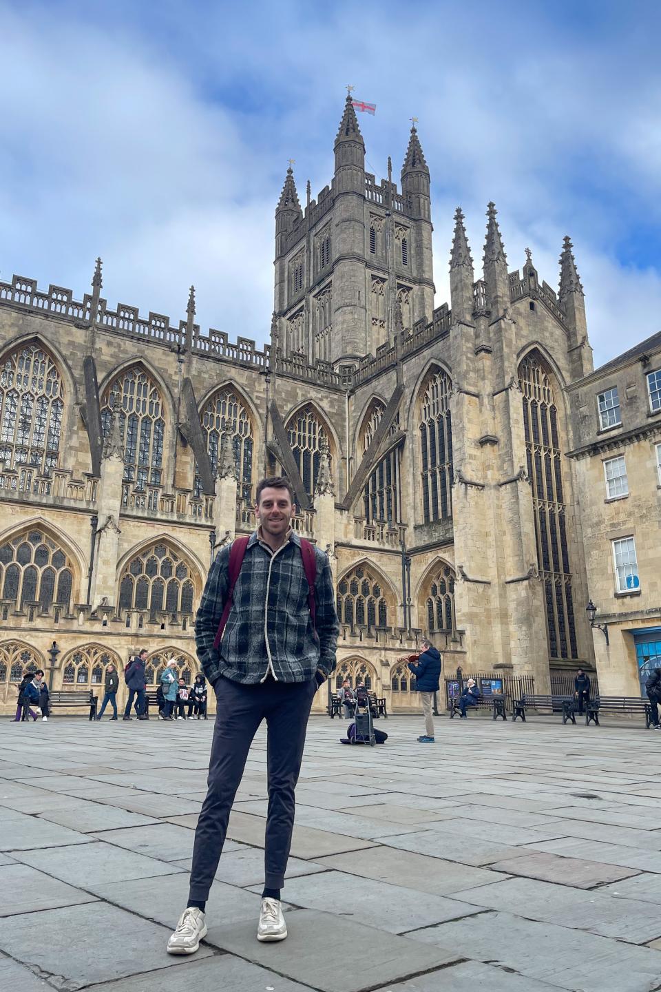 Man standing in front of Bath Abbey. Others are seen walking around the square