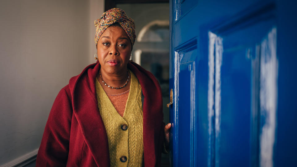 Michelle Grennidge in a red overcoat and green cardigan opens a blue door