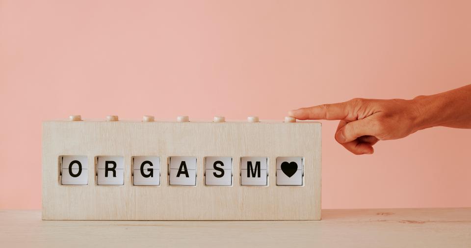 man pushing the button of a split flap display, with the text orgasm, on a wooden surface against a pale pink background, in a panoramic web banner format