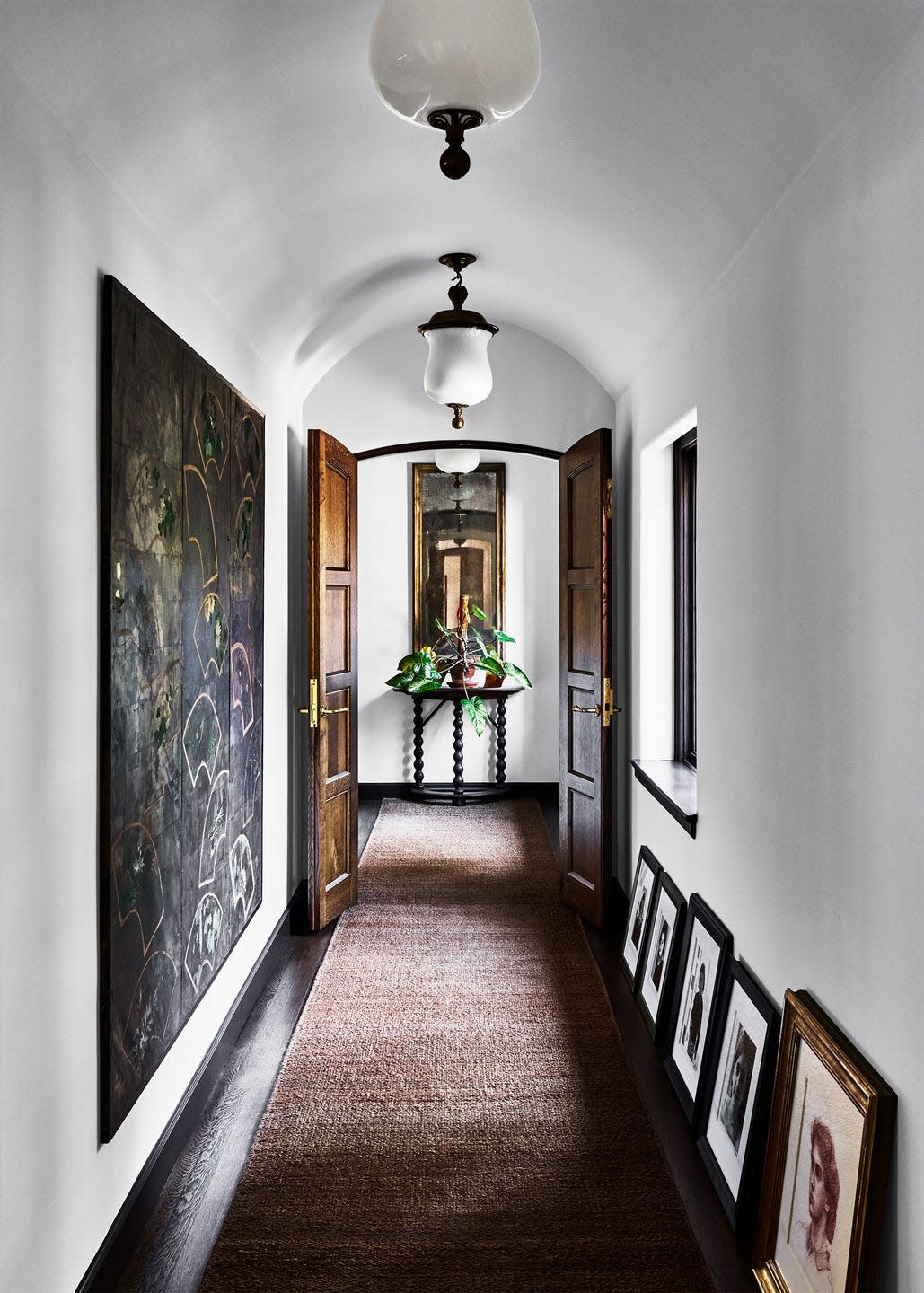 a hallway painted white with an arched ceiling, white pendants with wrought iron base, carpet runner, large artwork on wall and framed photos leaning against facing wall, open double wood doors, console table