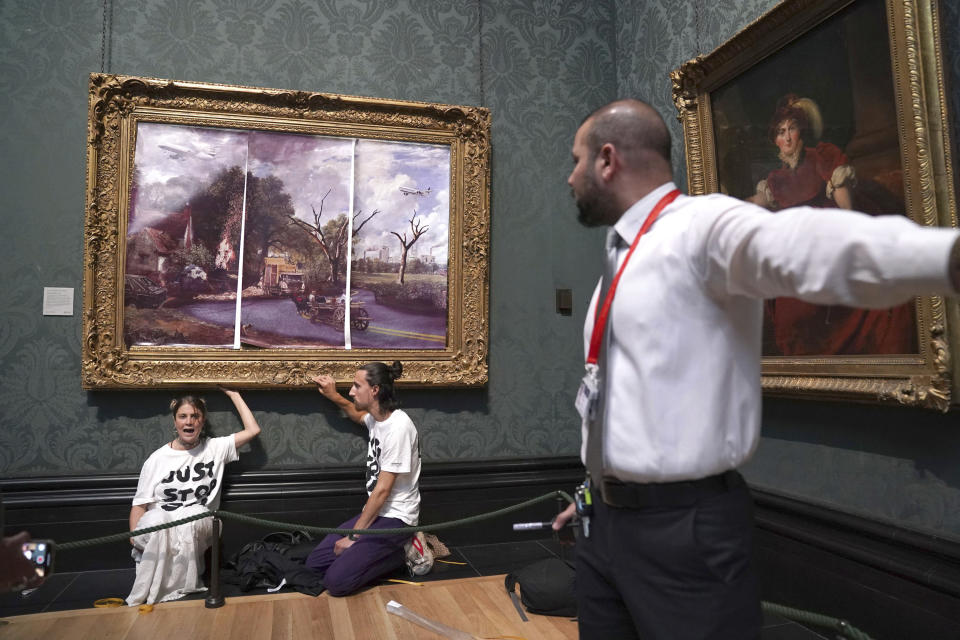 A security guard looks at protesters who glued their hands to the frame of John Constable's The Hay Wain, inside the National Gallery, London, Monday July 4, 2022. Police say two climate change protesters have been arrested after they glued themselves to the frame of a famous John Constable painting hanging in Britain’s National Gallery. The two, from the protest group “Just Stop Oil,” stepped over a rope barrier and covered “The Hay Wain” on Monday with large sheets of paper depicting “an apocalyptic vision of the future” of the landscape. (Kirsty O'Connor/PA via AP)
