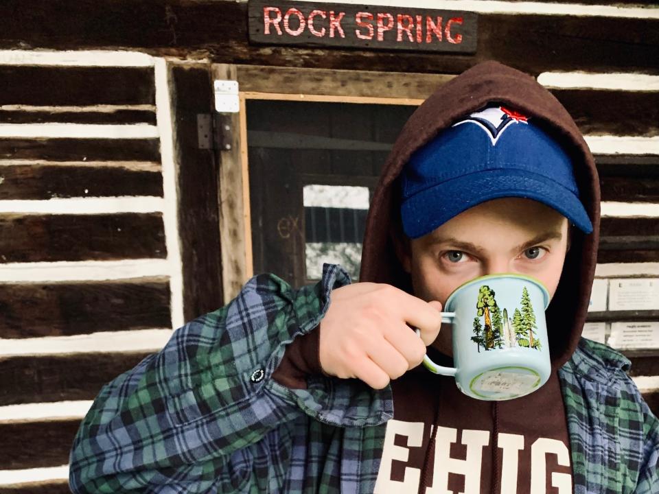 man drinking a cup of coffee in front of a cabin the has Rock Spring over the door.