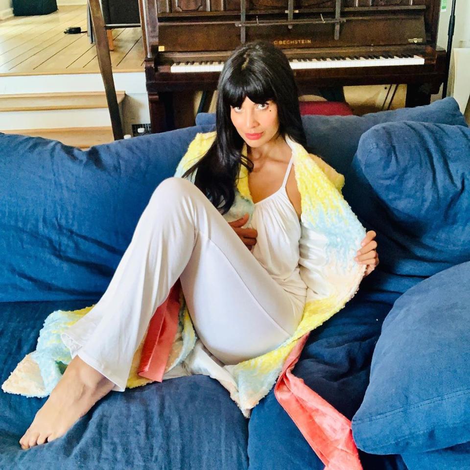 Jameela Jamil wearing white pajamas while sitting on her couch  to celebrate the Emmys 2020 from her home