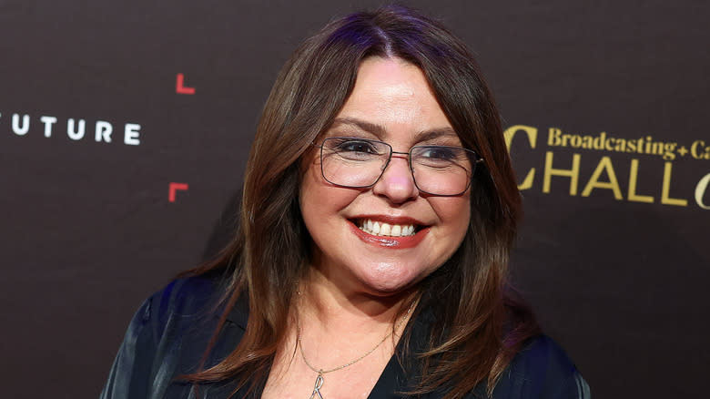 Rachael Ray wearing glasses and blue suit in front of black background