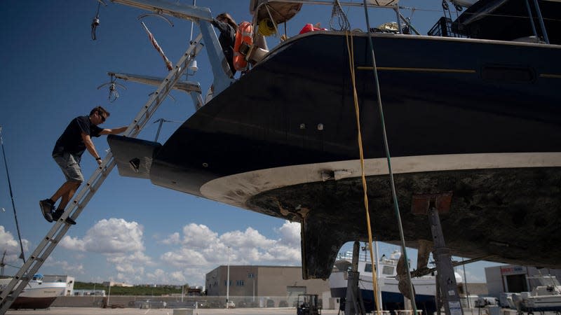 A man climbs down a ladder leading to a damaged sailing yacht sitting in a paved lot waiting for repairs. 