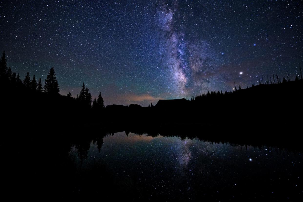 A starry night sky with milky way over a pond surrounded by forest.