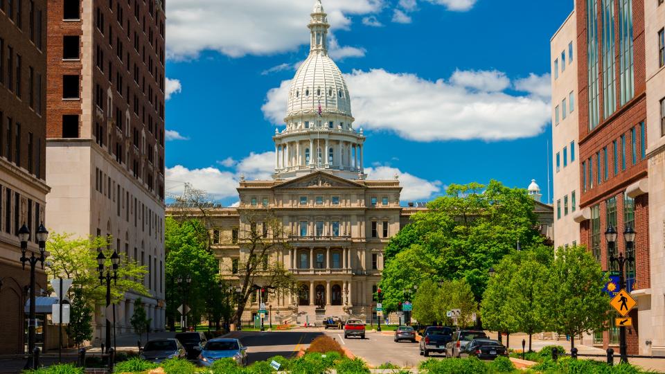 Lansing, United States - May 24, 2014 - The Michigan State Capitol as viewed from within Downtown Lansing, with trees, plants, office buildings, and pedestrians and cars and a driver in the foreground, and a blue sky with clouds in the background.