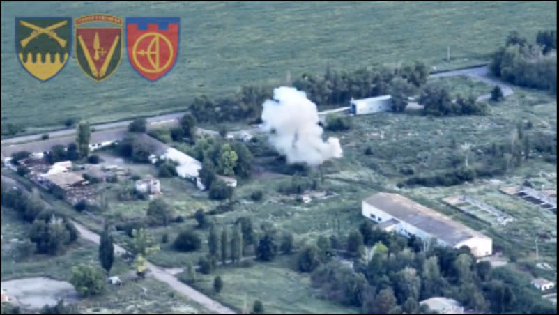 Grainy aerial image of several buildings and trees among a few roads and a grassy field with a large cloud of white smoke in the center.