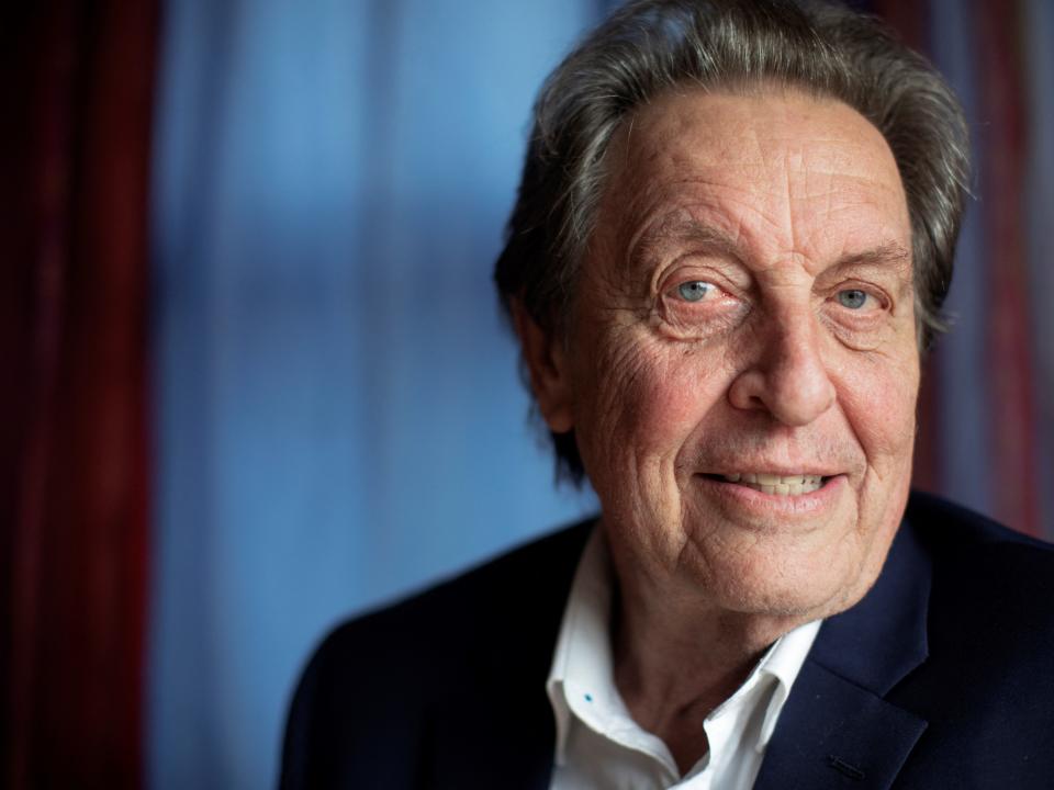 Errol Musk smiling while wearing a white shirt with the top button undone and a black blazer in a dimly-lit room.